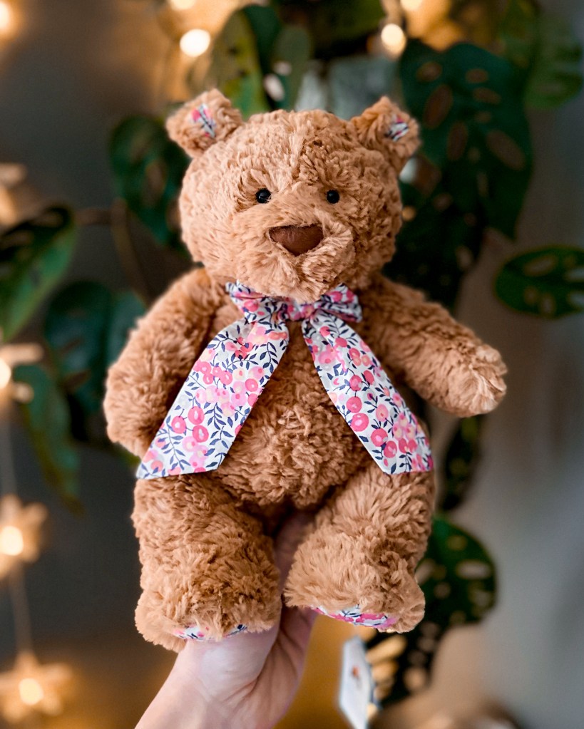 A medium Jellycat Bartholomew Bear wearing Liberty Wiltshire Berry print bow, held up in front of fairy lights and a Monstera plant — Enfys, the special edition Liberty bear featured in this blog post.
