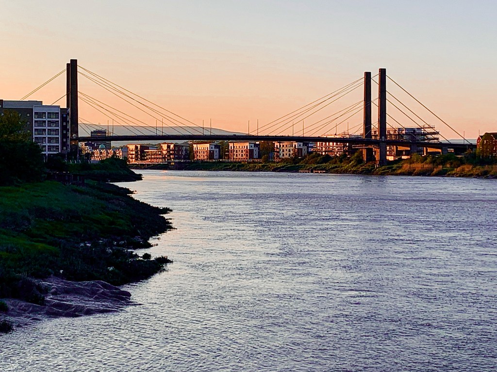 A picture of a bridge across a calm river in Wales near sunset, the orange glow reflecting on the bridge and buildings nearby and a mountain in the far background.