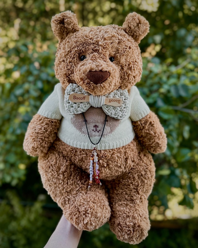 A Huge brown Jellycat bear wearing a cream bear jumper and macrame bow with the name Biscoff on it
